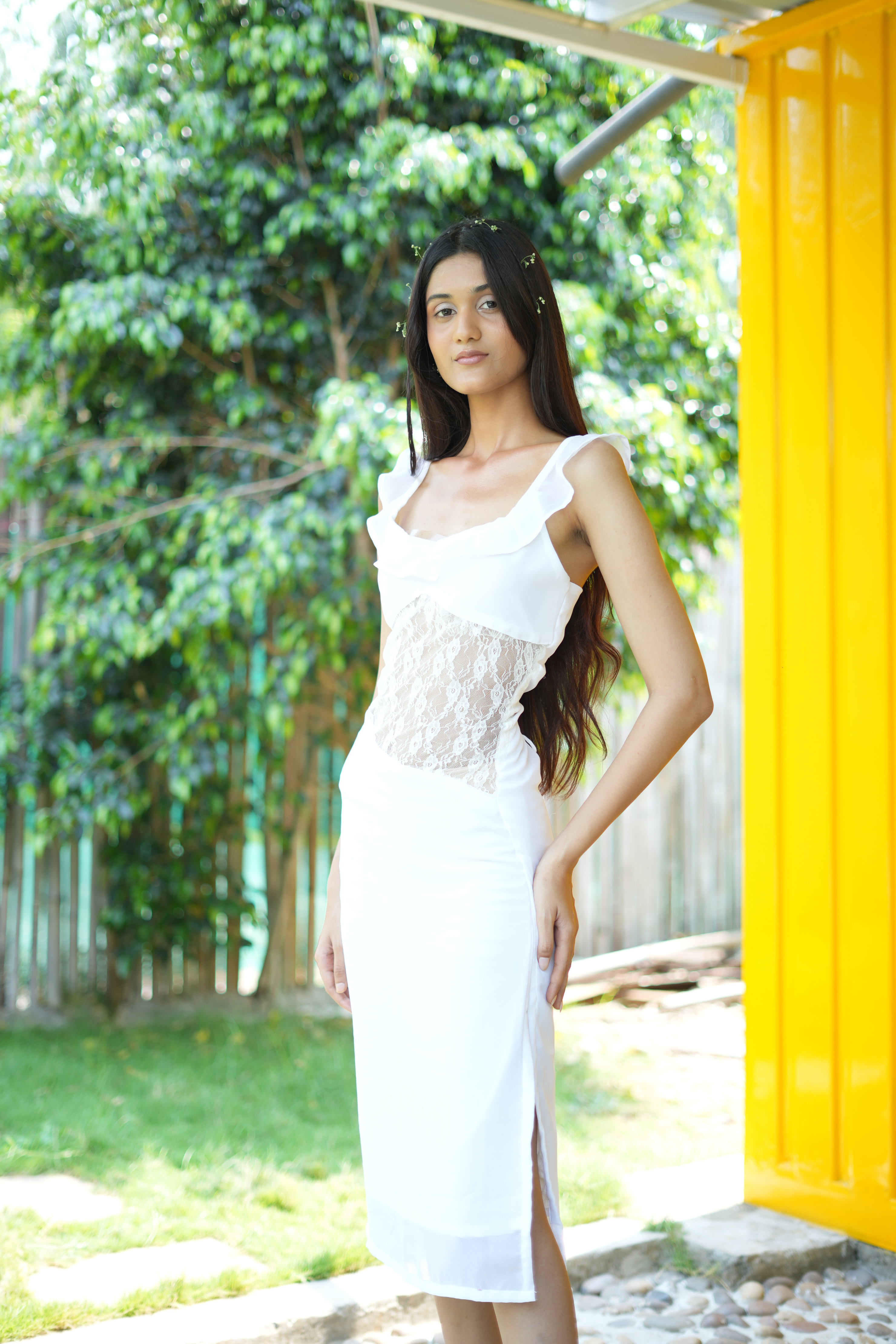 Woman in a white dress standing outdoors with greenery and a yellow curtain in the background